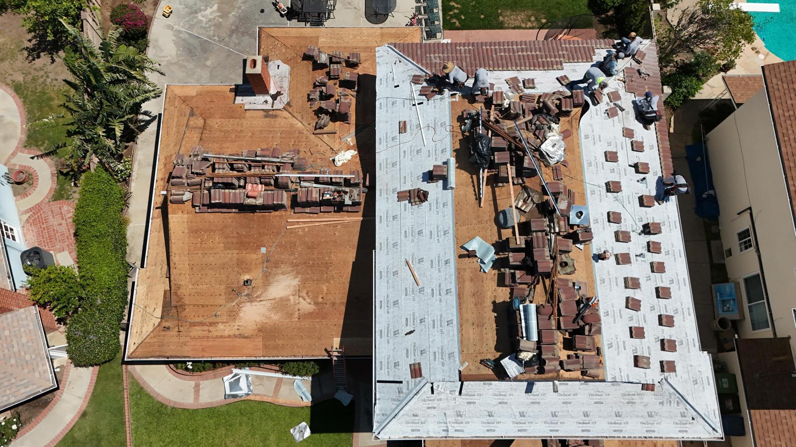 Aerial view of A&P re-roofing a two-section tile roof, crew laying underlayment and stacking clay tiles across the ridge